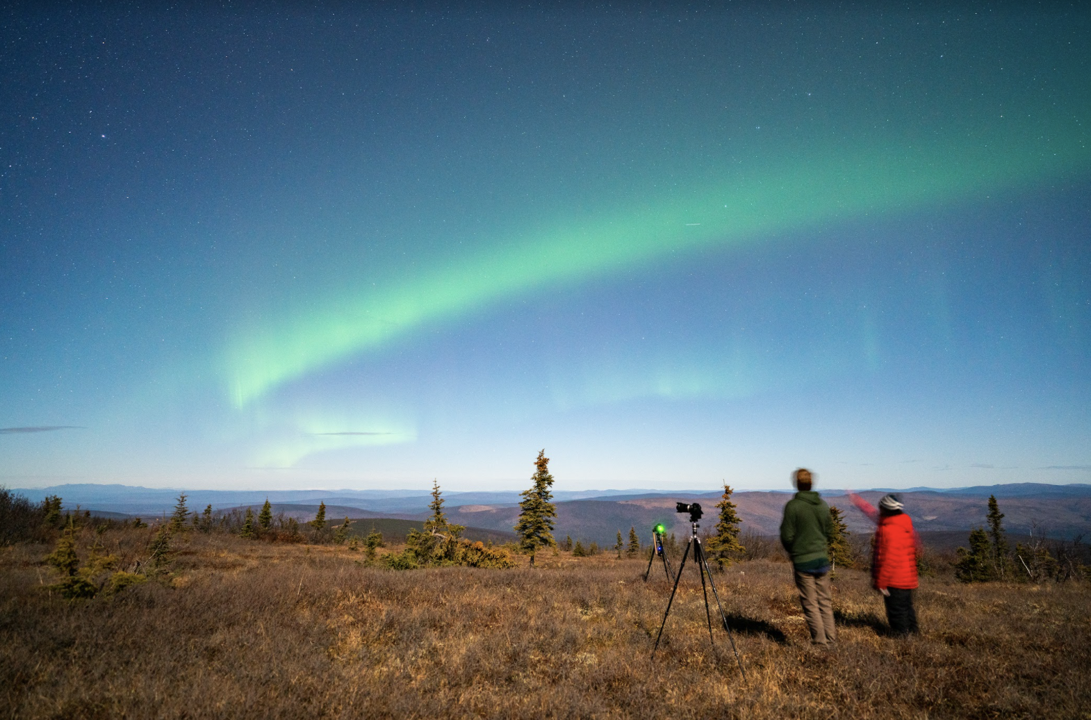 Two young men stand next to a camera looking up at waving green aurora.
